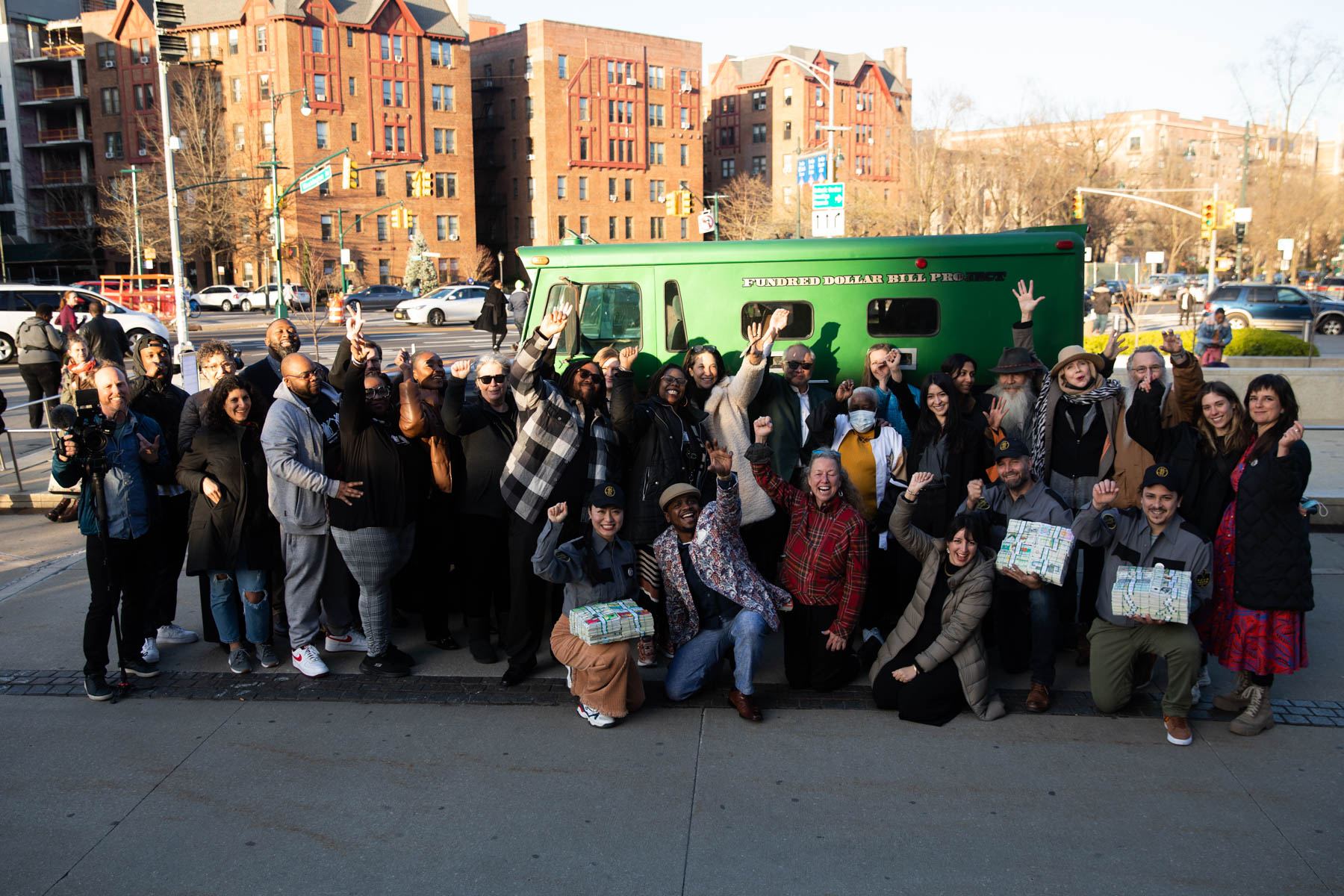 About 30 people outdoors standing and crouching in front of a green armored truck. The street, passing cars, pedestrians and residential buildings are pictured in the background.
