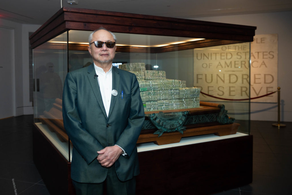 A person indoors standing in front of a glass display containing stacks of Fundred Dollar Bills. In the background, a plaque reads “BY THE PEOPLE OF THE UNITED STATES OF AMERICA FUNDRED RESERVE.”