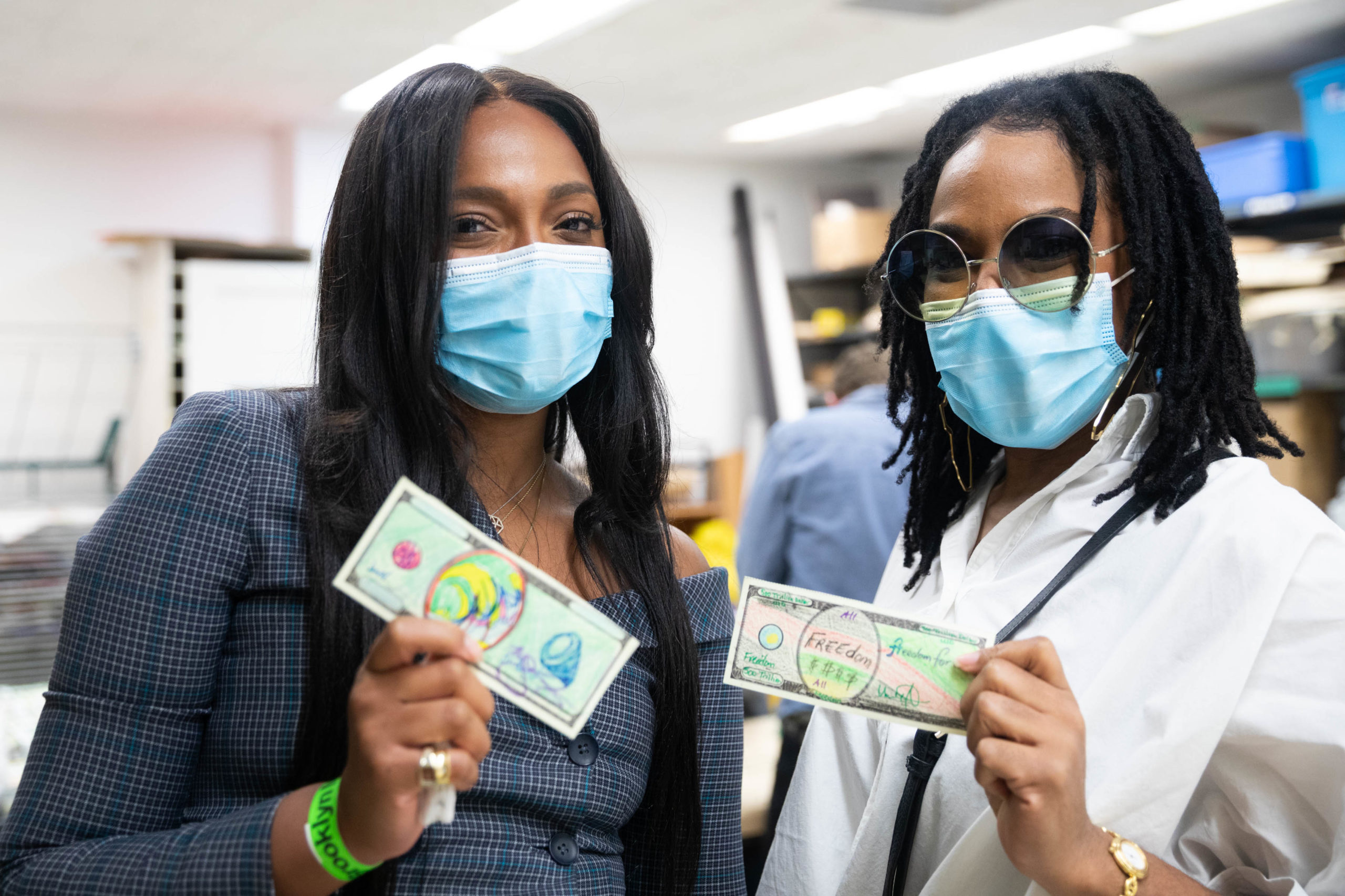 Two people indoors wearing masks and holding up Fundred Dollar Bills.